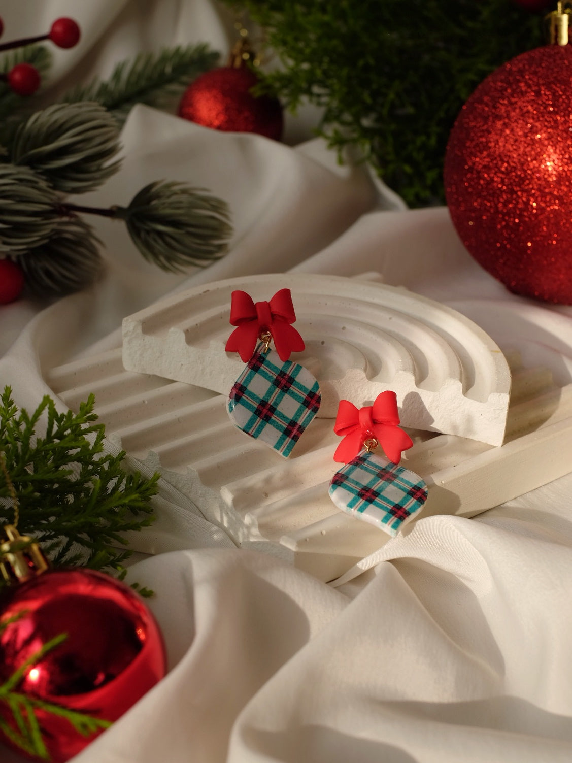 Pair of earrings with plaid design and red bows on a decorative stand with Christmas decorations.