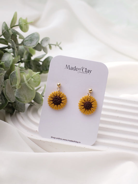 A pair of sunflower earrings made of polymer clay, displayed on a card with 'MADE OF CLAY' printed on it, placed on a white surface with a green plant in the background.